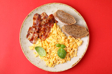 Delicious scrambled eggs with bacon and basil in plate on red background, top view