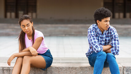 Two African American teenagers sit separately on steps outside. The girl is looking down, and guy is looking away from her. They appear to be having a disagreement.