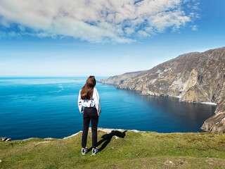Fototapeta premium Young teenager girl exploring beautiful Sliabh Liag cliff, county Donegal, Ireland. Travel and tourism. Warm sunny day. Stunning Irish nature landscape scene in the background.