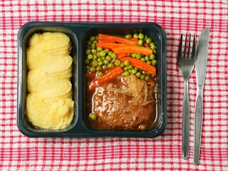 A meal consisting of burger, mashed potatoes and vegetables in plastic tray is served on a red and white checkered tablecloth and wooden table. Simple classic dish for take away. Office lunch.