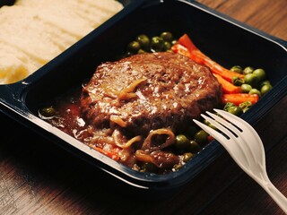 Cooked burger with vegetables in a black plastic tray on wooden table background and plastic fork. Part of ready meal for take away. Office lunch. Classic dish.
