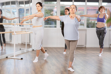 Poised senior woman, dressed in comfortable t-shirt and leggings, executing dance moves with focused precision among group of younger female ballet amateurs rehearsing in studio..