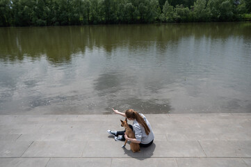 A young Caucasian woman and a cute little dog are walking in matching sneakers on the river bank. 