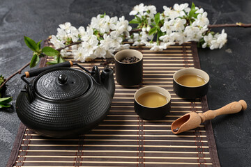 Teapot with cups, wooden spoon of dry tea and flowers on black grunge background