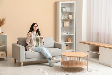Fototapeta premium Pretty young woman with cup sitting on grey armchair in stylish living room