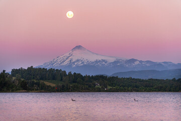 view of the Villarrica volcano reflected in the lake, with the full moon shining during a stunning...