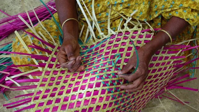 An old woman is making a colorful hand fan out of palm leaf fibers. Handicrafts are made by weaving colorful dry leaves.