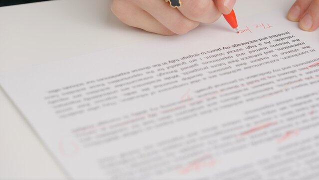 Professional school or college teacher grades a homework exam papers of students. Woman checks essay, crosses wrong words, corrects it with red pen.
