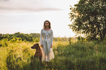 young cheerful woman playing with her dog outdoors in beautiful countryside field. Female and ridgeback dog walking and having fun