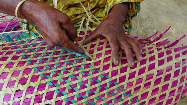 An old woman is making a colorful hand fan out of palm leaf fibers. Handicrafts are made by weaving colorful dry leaves.