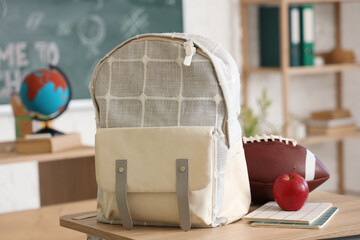 School desk with apple, backpack, football ball and notebook in light classroom, closeup