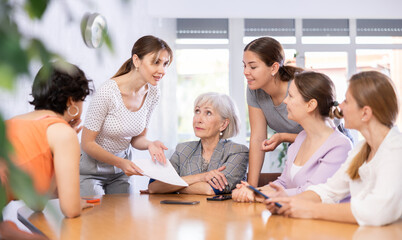 Fototapeta premium Cheerful young woman friendly talking to mixed age group of female colleagues sitting around table in office during informal meeting, discussing abstract topics or work-related situations