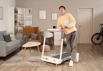 Young man with laptop near treadmill at home