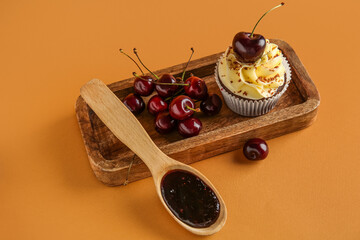 Wooden board with sweet cherry cupcake and spoon of jam on brown background