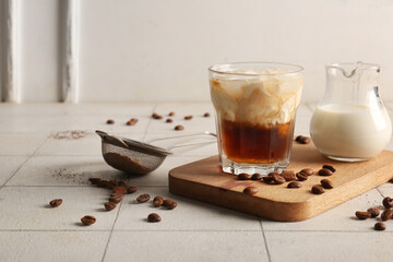 Glass of White Russian cocktail with coffee beans and jug of cream on white tile table