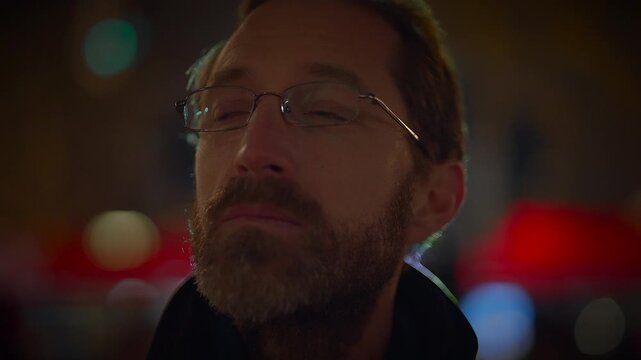 A man with glasses lost in thoughts under city lights and reflections in an urban setting at night