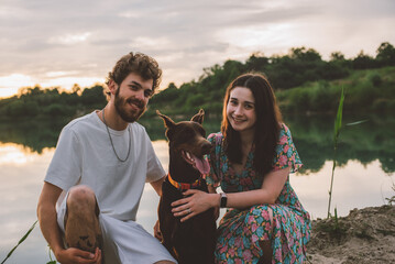 young happy couple having picnic and playing with their doberman dog outdoors near the lake during beautiful sunset