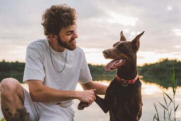 young man playing with his cute doberman dog outside near the lake during beautiful sunset 