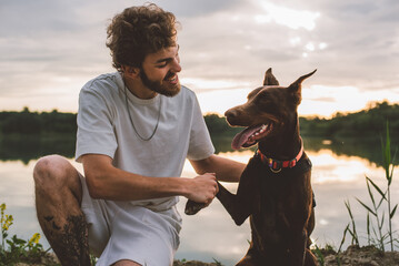 young man playing with his cute doberman dog outside near the lake during beautiful sunset 
