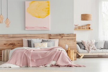 white and pink bedroom with wooden headboard, light grey floor, large pastel yellow abstract painting on the wall above bed frame, wood log stack side table, gray sofa in corner of room