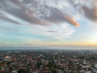 A serene dawn panorama over Yogyakarta, where tranquil clouds float above the sprawling cityscape, blending with the horizon to herald a peaceful start to the day.