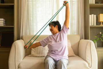 Happy Asian senior mature woman enjoy healthy lifestyle resistance band training exercise for stretching body muscle shoulder and arm on sofa in living room. Elderly self-care and home fitness concept
