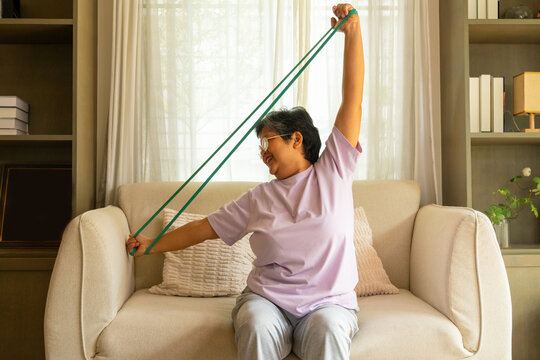 Happy Asian senior mature woman enjoy healthy lifestyle resistance band training exercise for stretching body muscle shoulder and arm on sofa in living room. Elderly self-care and home fitness concept
