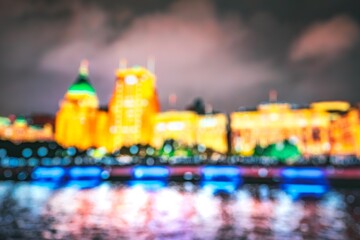A blurred view of the Shanghai skyline at night. The Bund and other buildings are lit up, reflecting in the water below.