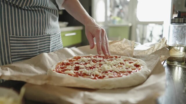 A female chef in an apron is putting mozzarella cheese on a raw pizza. Cooking delicious pizza at home in the kitchen.