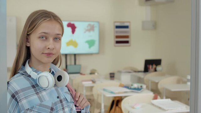 Portrait of teenage blonde Caucasian girl with long hair and blue eyes standing in classrom during recess, smiling and looking at camera