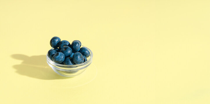 Fresh Blueberries in a glass bowl on a light yellow background, minimal summer fruits concept, banner with copy space