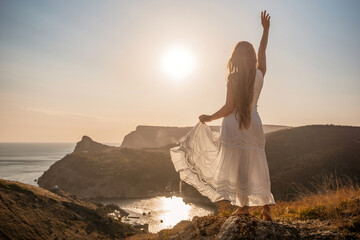 A woman in a white dress stands on a rocky hill overlooking the ocean. She is smiling and she is happy.