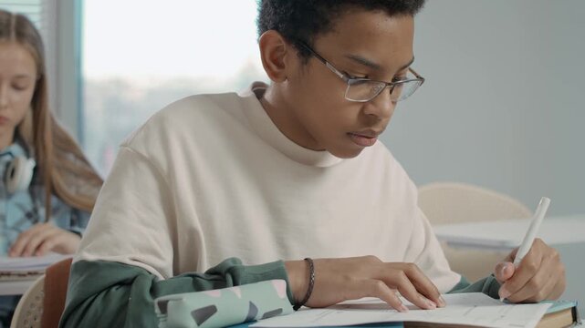 Close-up of teenage left-handed African American boy sitting at desk, thinking and writing with pen while completing challenging task during lesson