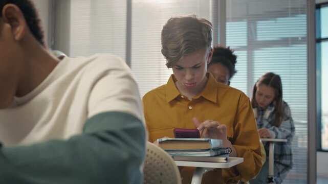 Close-up of cheerful Caucasian teenage boy sitting at desk among diverse classmates at school, cheating at test by using Internet on smartphone during lesson