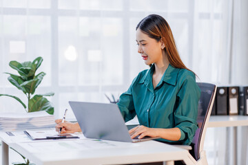 Asian woman in the workplace Work Hard writing notes on a pad from her laptop computer with analytical charts and graphs in front of her, business people concept