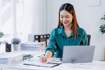 Asian woman in the workplace Work Hard writing notes on a pad from her laptop computer with analytical charts and graphs in front of her, business people concept