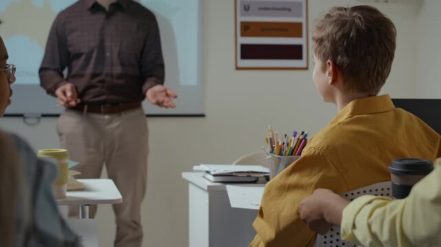 Back view of talkative teenage Caucasian boy sitting at desk in classroom distracting classmates by chatting and interrupting course of lesson