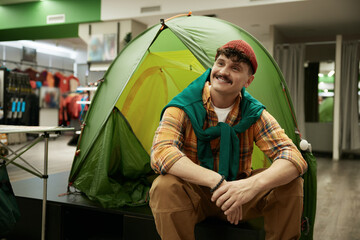 Happy smiling hipster man bicyclist sitting nearby travel camp tent in store