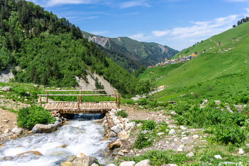 A clear mountain river in a stone bed among grass and yellow flowers through the stones. Wooden bridge with railings and mountains in the background