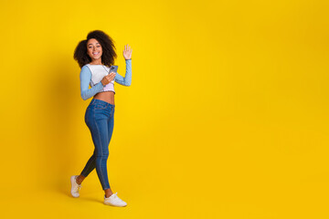 Full body photo of attractive young woman hold device walking wave hand dressed stylish blue clothes isolated on yellow color background