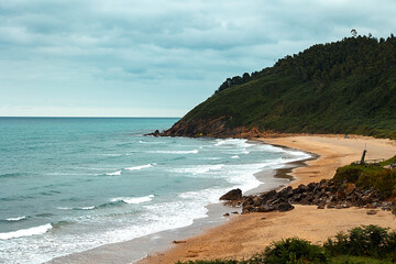Sandy beach with gentle waves and green hillside under a cloudy sky
