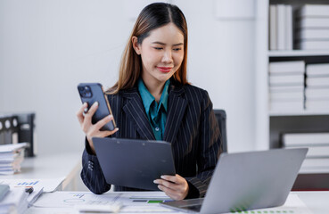 Asian woman sit at workplace desk holds cellphone staring at laptop, synchronize data between...