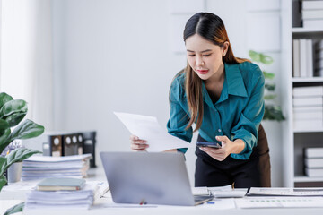 Asian woman sit at workplace desk holds cellphone staring at laptop, synchronize data between...