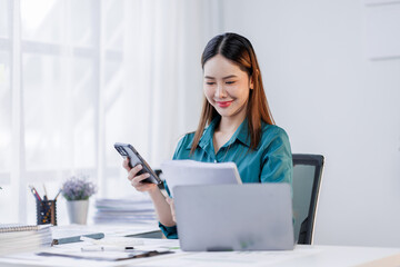 Asian woman sit at workplace desk holds cellphone staring at laptop, synchronize data between...