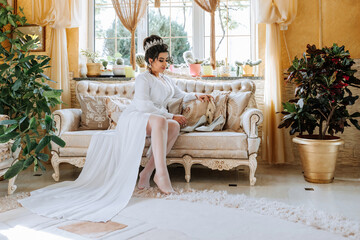 A woman in a white dress sits on a couch in a living room. She is wearing white stockings and a tiara. The room is decorated with potted plants and a vase