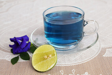 Purple hot water in a glass cup with saucer Lemon wedge and purple Clitoria ternatea flowers with leaves All placed on a table with a plastic stencil with a crab pattern on it.

