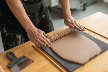 Close-up of a potter's hands rolling out clay using a rolling pin with patterns. 