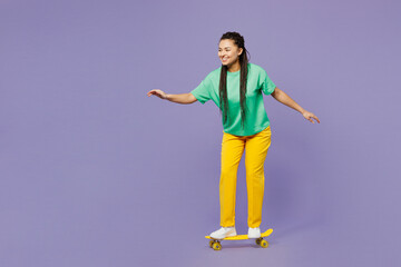 Full body side profile view young woman of African American ethnicity with dreadlocks wear t-shirt casual clothes riding skateboard pennyboard isolated on plain purple background. Lifestyle concept.