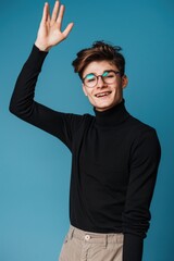 Naklejka premium A young man with glasses, waving at the camera. He is dressed in a black turtleneck and beige pants.