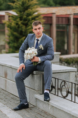 Obraz premium A man in a suit and tie sits on a stone wall holding a bouquet of flowers. Concept of elegance and sophistication, as the man is dressed in formal attire